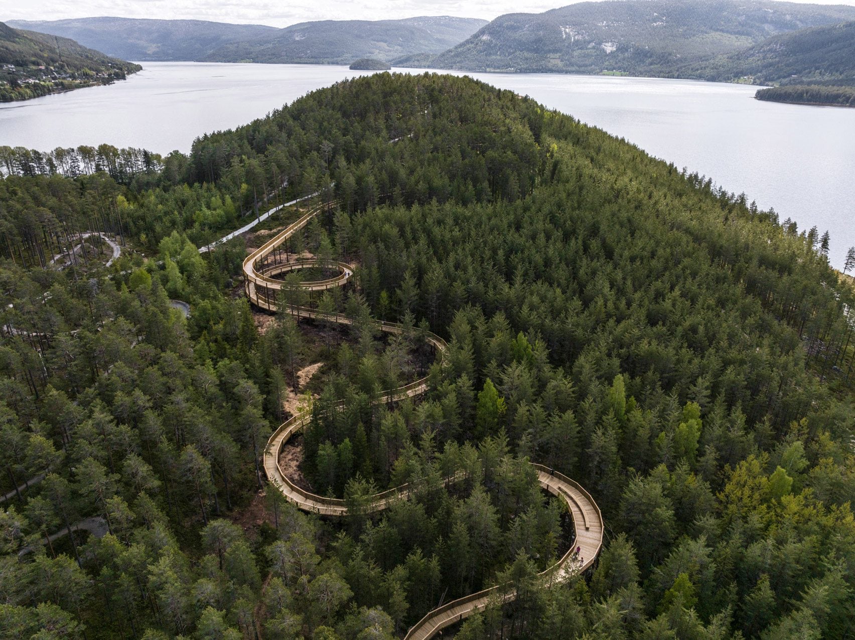 Une passerelle accessible dans les arbres à travers le parc Hamaren en Norvège 21 Photo aérienne d'une promenade sinueuse en pin au milieu d'une forêt près d'un lac et d'une colline escarpée.