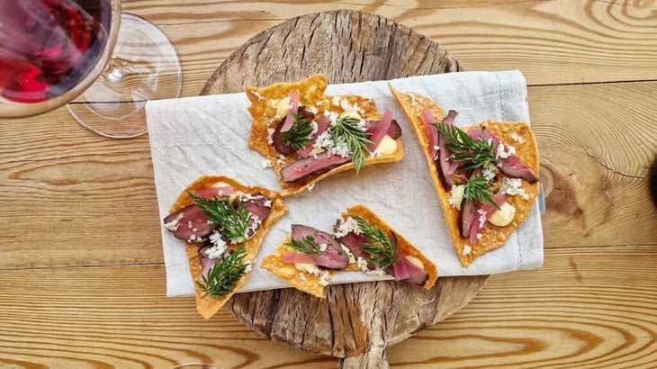 Voici les meilleures cabanes côtières de Norvège 22 Un plateau circulaire en bois avec des crudités de cœur de bœuf grillé sur des crackers de pois jaunes, avec un verre de vin rouge à gauche.