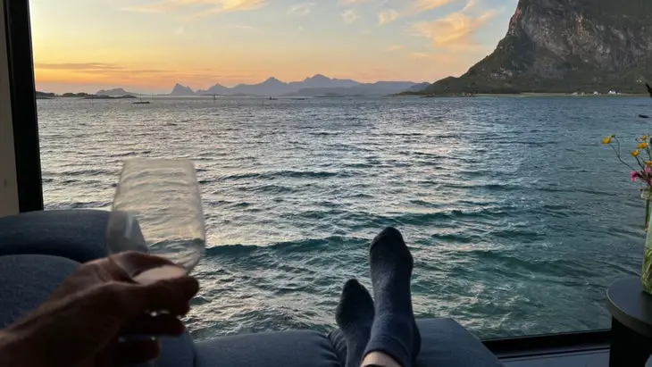 Vue d'un coucher de soleil aux couleurs pastel sur le front de mer et les montagnes. On peut voir une personne se détendre avec des chaussettes et un verre de vin à la main.