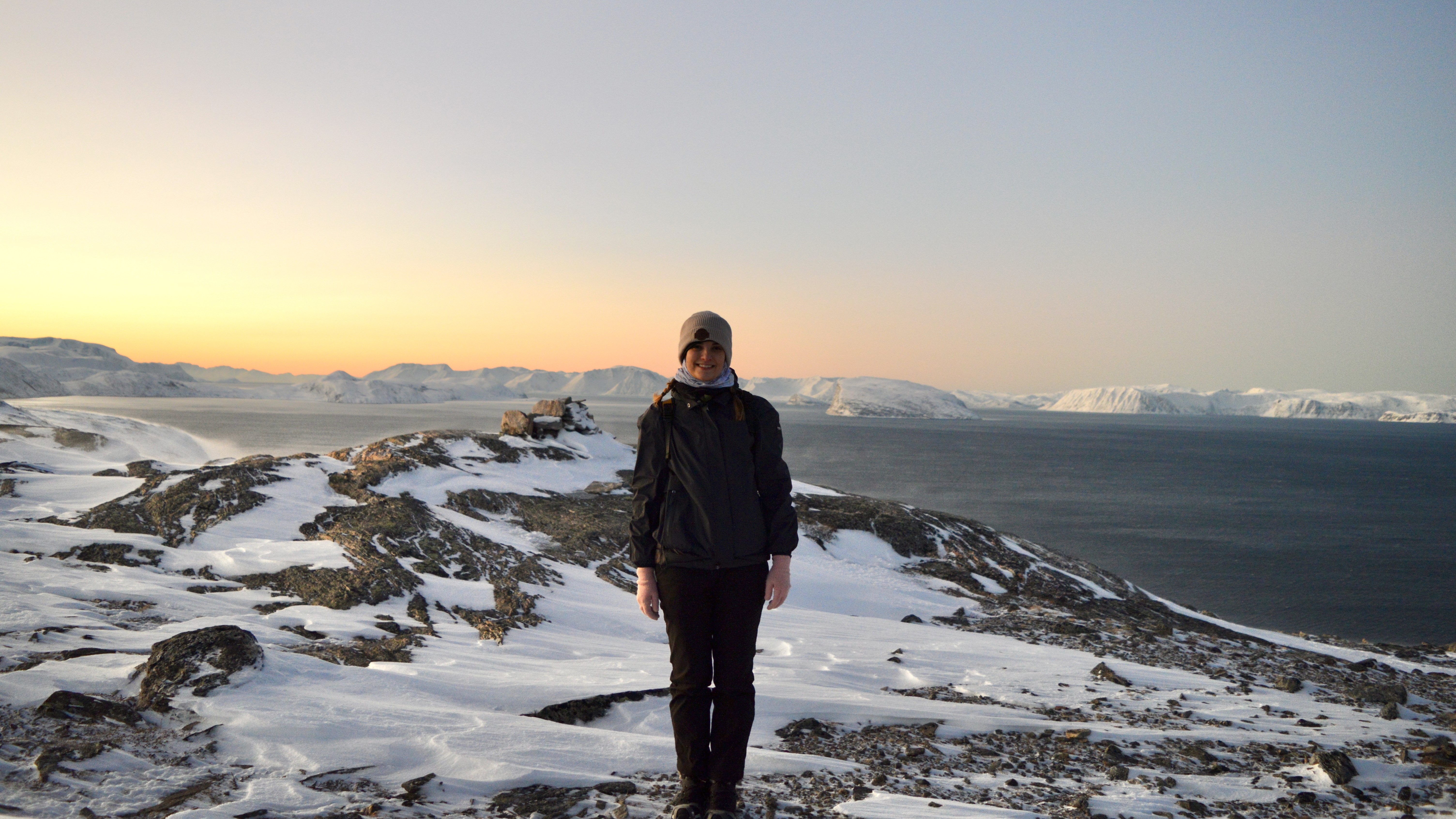 Une femme se tient au centre de l'image, avec une montagne enneigée sous les pieds et la mer au loin.