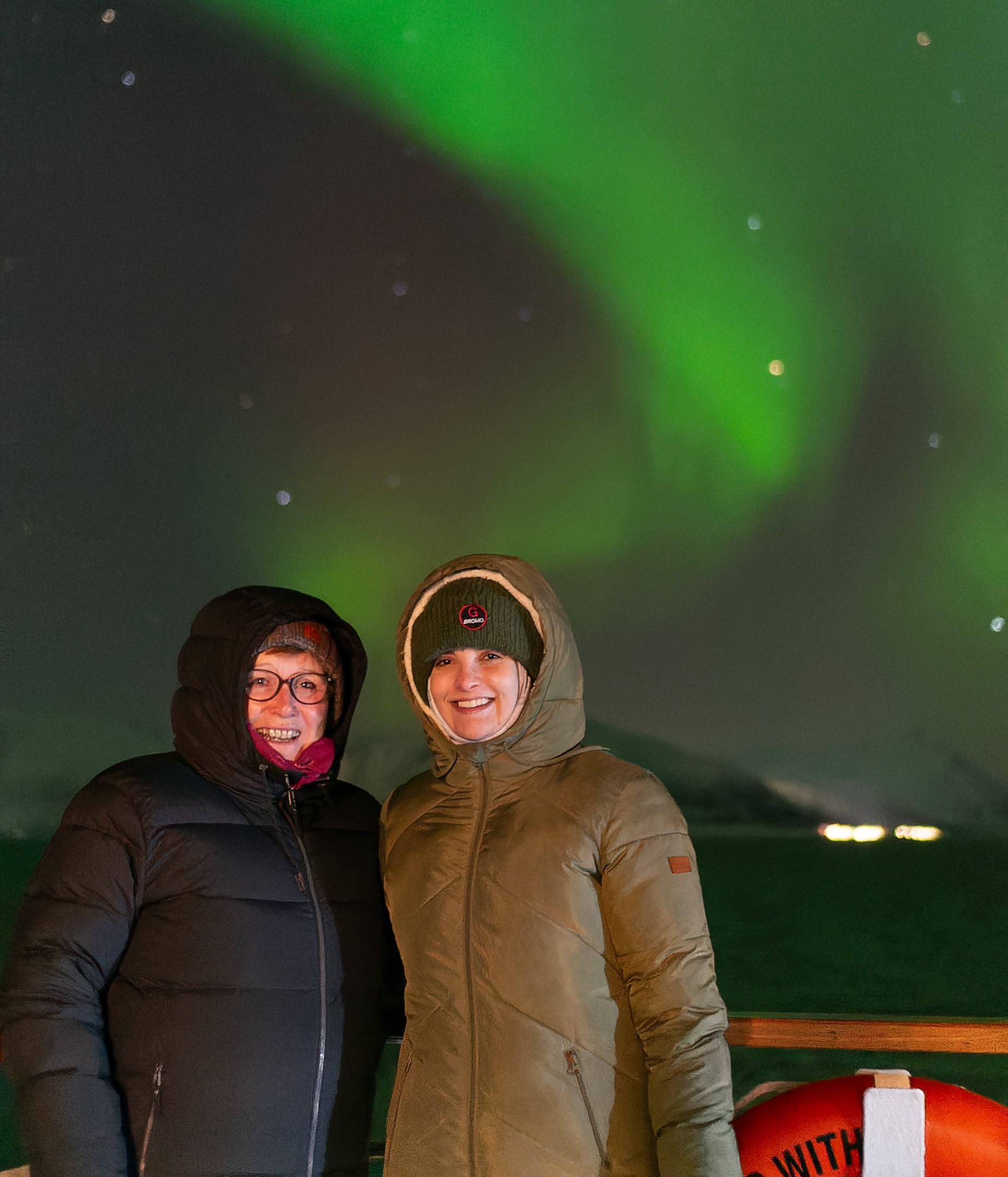 Deux femmes vêtues de longs manteaux et souriantes avec une aurore boréale derrière elles.