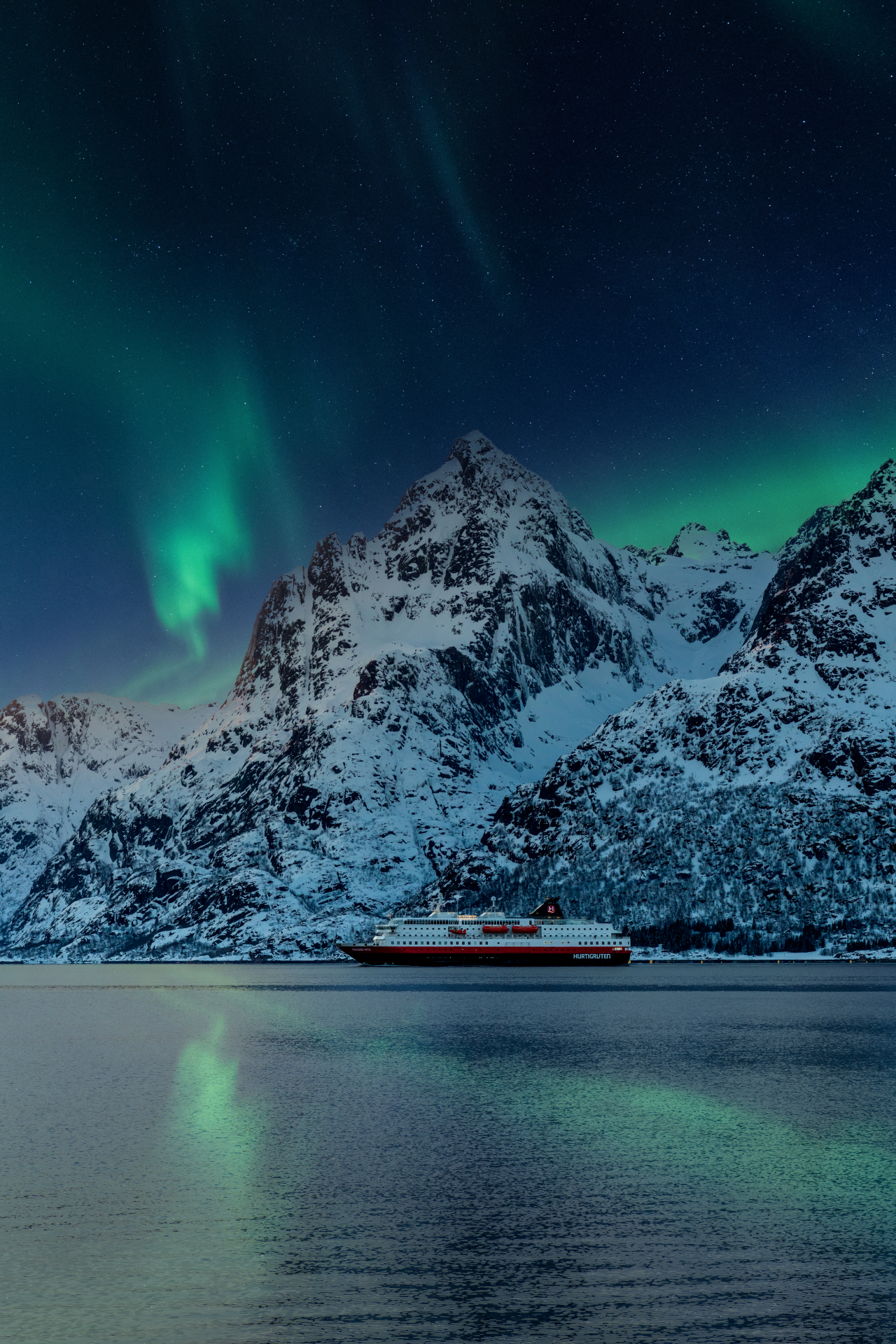 Le navire Hurtigruten sous une aurore boréale avec de hautes montagnes en arrière-plan.