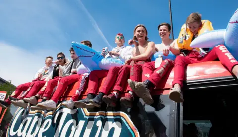 Shutterstock Un groupe d'élèves en bleu de travail est assis dans un bus russe rouge, souriant et buvant, avec des jouets de piscine gonflables, dans un ciel bleu clair, avec les mots 
