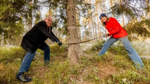 Sturlason/Oslo kommune Les deux maires se tiennent de part et d'autre d'un tronc d'arbre avec une scie à deux hommes. Ils regardent la caméra et sourient en s'appuyant sur leurs jambes. Le maire Anne Lindboe porte un chapeau et un manteau orange et le maire Paul Dimoldenberg porte un manteau noir et un jean bleu.